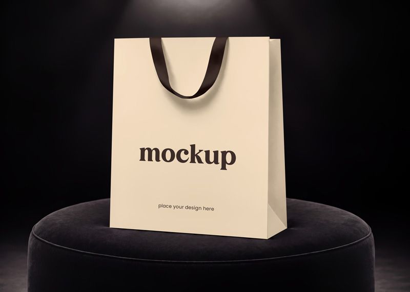 Paper shopping bag mockup with minimalist logo and dark ribbon handles on a black studio pedestal