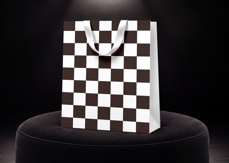 Paper shopping bag mockup with brown and white checkerboard pattern and white ribbon handles in a dark studio setting