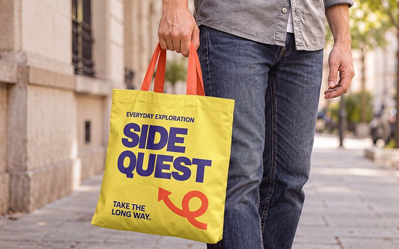 Canvas tote bag mockup in hand with yellow fabric, orange handles, and purple lettering on a city sidewalk