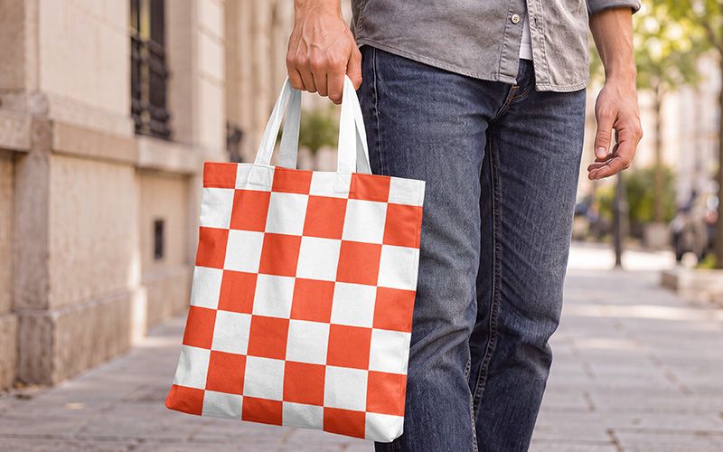 Canvas tote bag mockup in hand with an orange and white checkerboard print and white handles