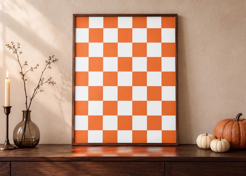 Autumn poster mockup with an orange and white checkerboard print in a wooden frame beside pumpkins and a candle