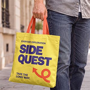 Yellow canvas tote bag mockup in hand with orange handles and bold purple lettering